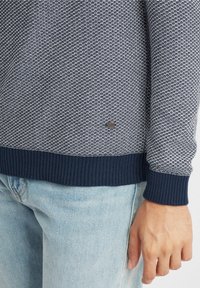 Pull en maille bleu marine avec un motif texturé gris clair. Bas et poignets côtelés. Petit logo de marque métallique sur le côté inférieur.