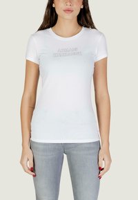 Vrouw die een nauwsluitend wit Armani Exchange T-shirt met korte mouwen en lichtgrijze jeans draagt, staand tegen een effen achtergrond.