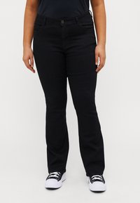 Svart jeans med rett ben-design, laget av denim. Har frontlommer og en enkel messingknapplukking. Brukt med hvite joggesko.