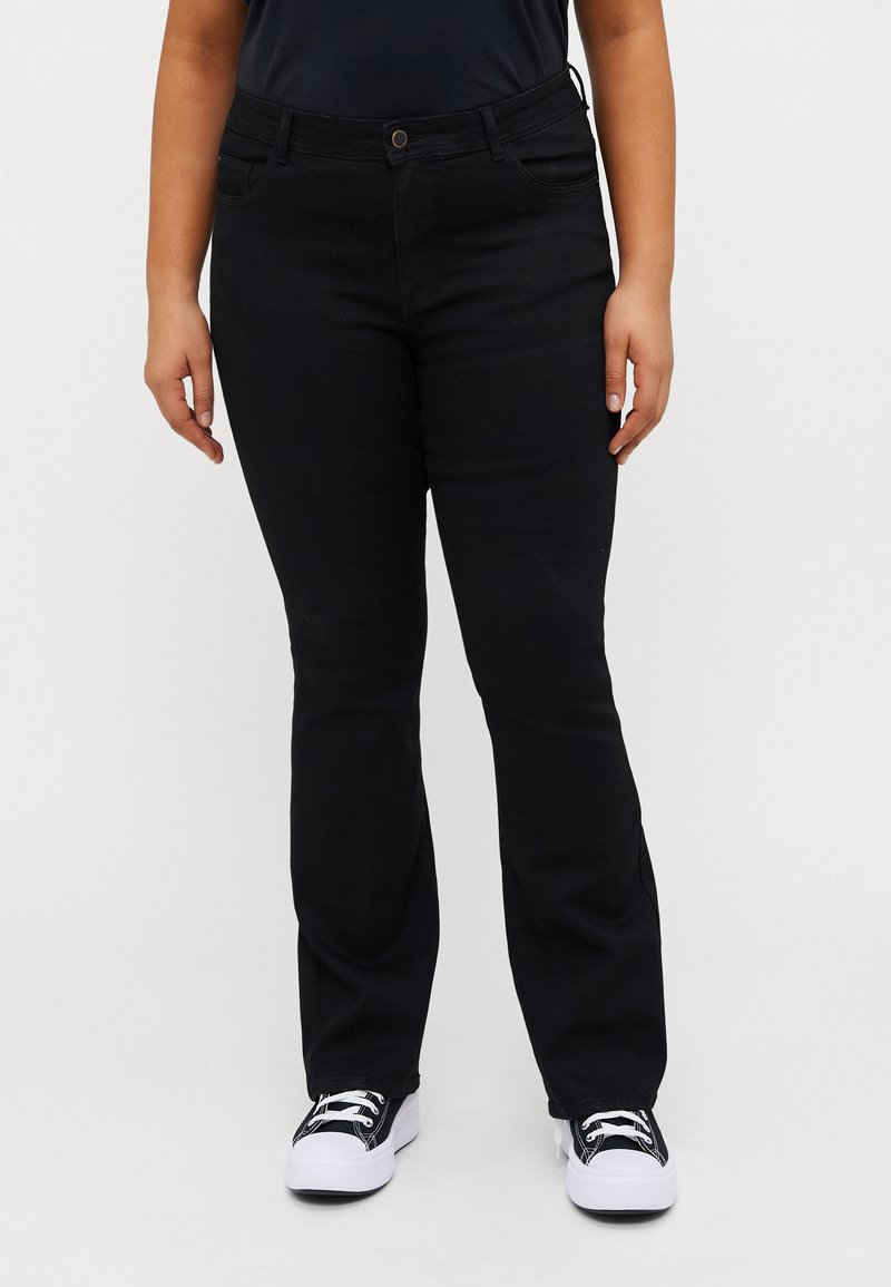 Svart jeans med rett ben-design, laget av denim. Har frontlommer og en enkel messingknapplukking. Brukt med hvite joggesko.