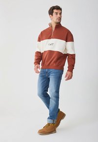 Ritsvest in brandoranje met een witte horizontale streep en een centraal logo. Gecombineerd met blauwe spijkerbroek en beige laarzen.