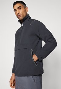 Donkergrijze quarter-zip jas met een hoge kraag, elastische manchetten, voorzak en subtiele reflecterende accenten. Glad, lichtgewicht materiaal.