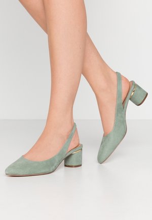 Nainen pukeutunut vihreisiin mokkanahkaisiin slingback-korkokenkiin, joissa on paksu korko ja kultaiset reunat, seisoo valkoisella lattialla yksivärisen taustan edessä.