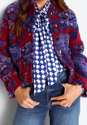 Nainen, joka pukeutunut punaisesta ja violetista koostuvaan kuvioituun kukkatakkiin sinivalkoisen pilkullisen rusetillisen paidan päällä, yhdistettynä sinisiin farkkuihin.