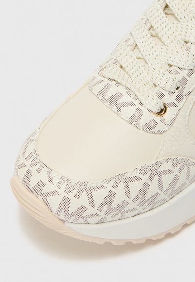 Zapatilla de color claro con un upper liso y texturizado que presenta un patrón geométrico. Punta redondeada, con una suela acolchada y diseño de cordones.