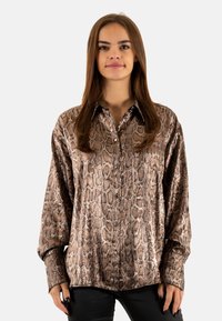 Camisa de manga larga de estampado de serpiente marrón con cuello clásico, cierre de botones en la parte delantera y una textura suave y brillante. Combinada con pantalones negros.