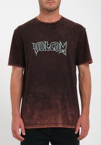 Mies, joka on pukeutunut haalistuneeseen tummanruskeaan t-paitaan, jossa rintaosassa on vaaleansiniset, sahalaitaiset "VOLCOM"-kirjaimet.