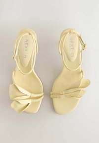 Sandales pour femmes couleur crème avec brides à la cheville et détail décoratif en cuir plié sur les orteils, vues de dessus sur un fond clair.
