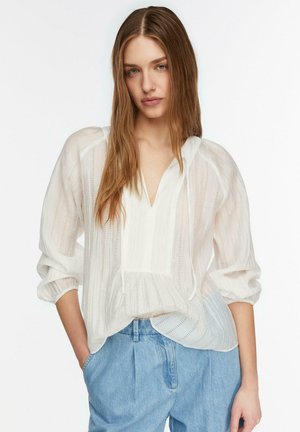 Kobieta o długich brązowych włosach, ubrana w białą teksturowaną bluzkę z luźnymi rękawami oraz jasnoniebieskie, wysokowytaliowane dżinsy, ręce w kieszeniach.