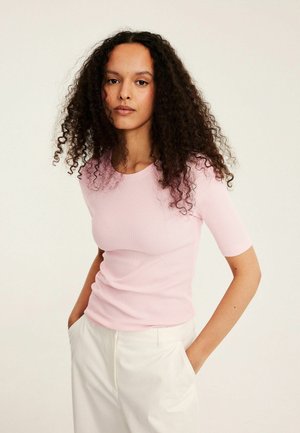 Vrouw met lang krullend haar die een lichtroze korte mouw top en een witte broek draagt, staand met handen in de zakken tegen een effen achtergrond.