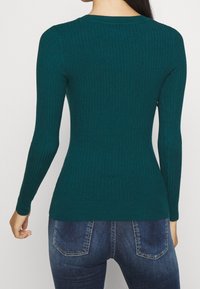 Dopasowany, turkusowy sweter z długim rękawem i prążkowaną strukturą, z okrągłym dekoltem i fakturowaną tkaniną. Pokazany od tyłu, noszony z ciemnoniebieskimi dżinsami.