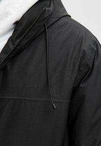 Črna lahka jakna z zadrgo spredaj, kapuco in nastavljivo vrvico. Gladka tekstura z mat površino in minimalnim okrasjem.