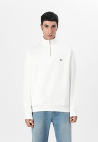 Vit zip-up hoodie med ribbad textur, hög krage och broderad grön logotyp. Kombination med ljusblå jeans.