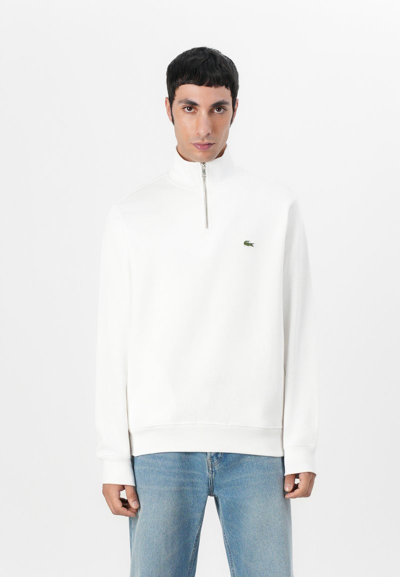 Vit zip-up hoodie med ribbad textur, hög krage och broderad grön logotyp. Kombination med ljusblå jeans.