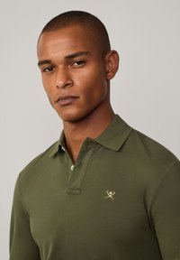 Maslinasto zeleni longsleeve polo majica s ovratnikom, koja ima mali zlatni grb i detalje s gumbima, izrađena od teksturiranog pamučnog materijala.