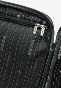 Close-up van de binnenkant van een zwarte stoffen koffer met een gestreepte voering, een mesh zak en twee zilveren metalen ritsen met het label "WITTCHEN."