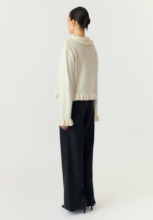 Donna con capelli raccolti in uno chignon che indossa un maglione crema con bordi arricciati e pantaloni neri a gamba larga, in piedi di lato su uno sfondo semplice.
