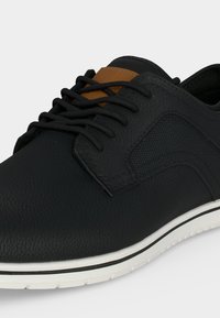 Chaussure décontractée noire à lacets avec panneau latéral texturé, languette marron et semelle blanche avec détail bande noire.