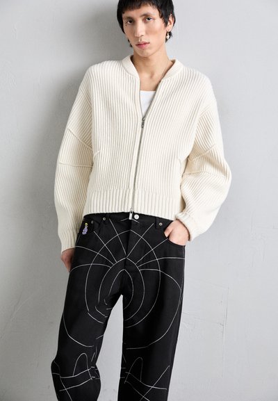 Cardigan crem cu torsade, cu fermoar, asortat cu pantaloni negri cu modele geometrice albe și un mic patch colorat pe buzunar.