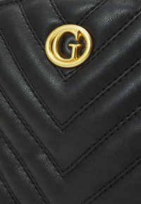 Borsa in pelle nera con un motivo a zig zag, cuciture dettagliate e un prominente accento dorato a forma di cerchio con una 'G' al centro.