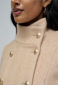 Mujer con un abrigo beige de espiga con botones redondos dorados y cuello alto, mostrando parte de su rostro y cabello negro.