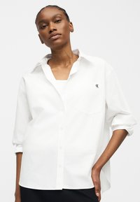 Νεαρή γυναίκα με πλεκτά μαλλιά, που φοράει λευκό, φαρδύ πουκάμισο με κουμπιά και γυρισμένα μανίκια και με μικρό λογότυπο CK στην τσέπη στο στήθος.