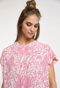 Blusa rosa con motivo paisley, maniche corte e scollo rotondo. Il tessuto sembra leggero e testurizzato, con un sottile dettaglio di bottoni sul davanti.