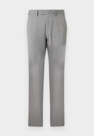 Mężczyzna stojący z rękami w kieszeniach, ubrany w granatową koszulę z długim rękawem, jasnoszare spodnie i czarne eleganckie buty na białym tle.