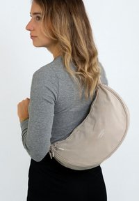 Borsa a spalla in tessuto beige con design curvo, dotata di chiusura a zip e un logo sottile. Texture liscia e aspetto minimalista.