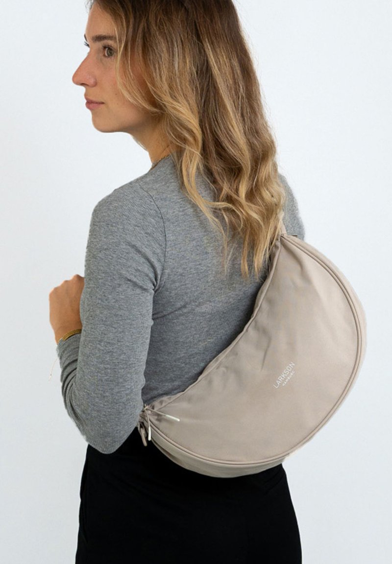 Borsa a spalla in tessuto beige con design curvo, dotata di chiusura a zip e un logo sottile. Texture liscia e aspetto minimalista.