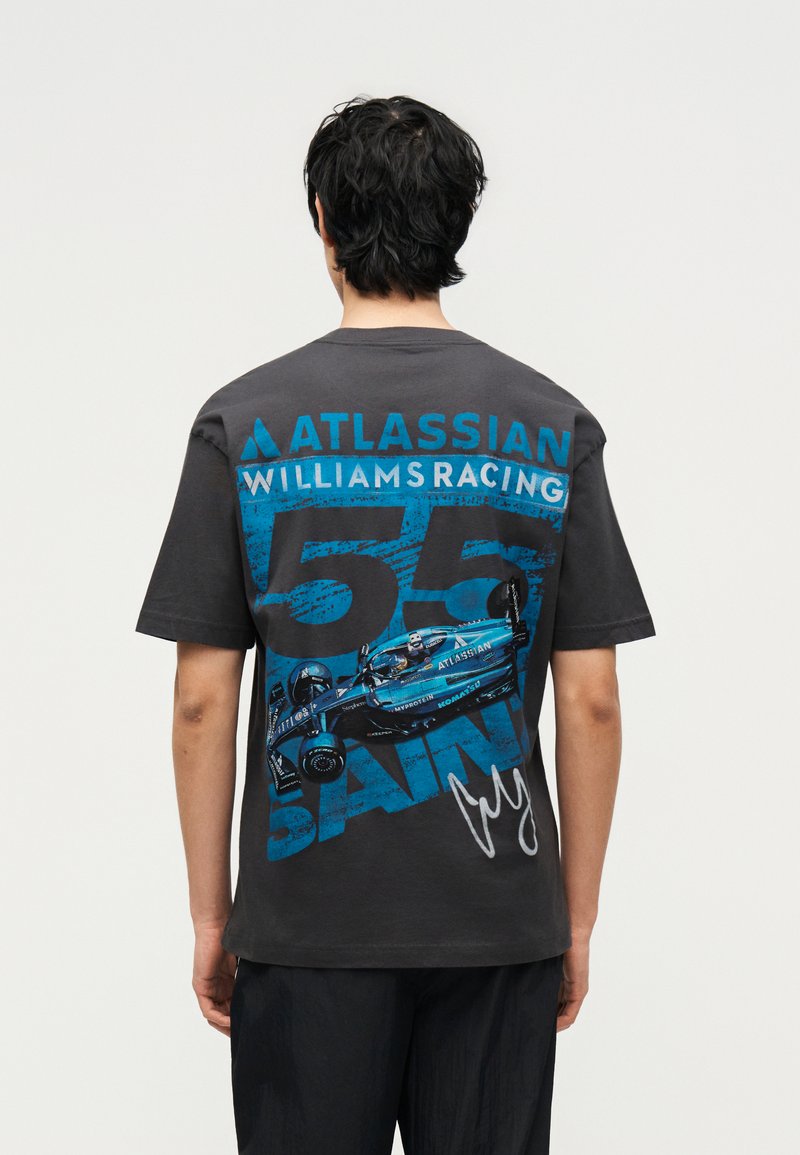 Άτομο με σκούρα μαλλιά που φοράει μαύρο μπλουζάκι με ένα μπλε Williams Racing Formula 1 αυτοκίνητο, αριθμός 63, και λογότυπα χορηγών στην πλάτη.