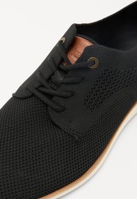 Juodas tinklinis batas su tekstūruotu viršumi, odiniu akcentu šalia liežuvėlio ir plokščiomis raišteliais. Padas baltas su beige kraštu.