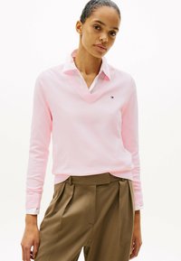 Hellrosa langärmliger Pullover mit V-Ausschnitt, über einem weißen Hemd mit Kragen getragen, kombiniert mit braunen hochtaillierten Hosen.