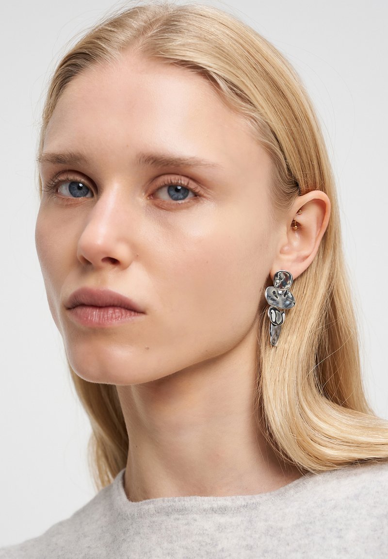 Orecchini astratti argentati con una finitura testurizzata, caratterizzati da un design fluido. Indossati da un modello con capelli biondi lisci e trucco minimale.