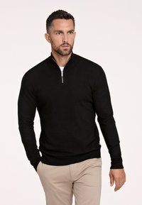 Maglione nero con collo zip, maniche lunghe, polsini e orlo a coste. Indossato sopra una camicia bianca, abbinato a pantaloni beige chiaro. Design pulito.