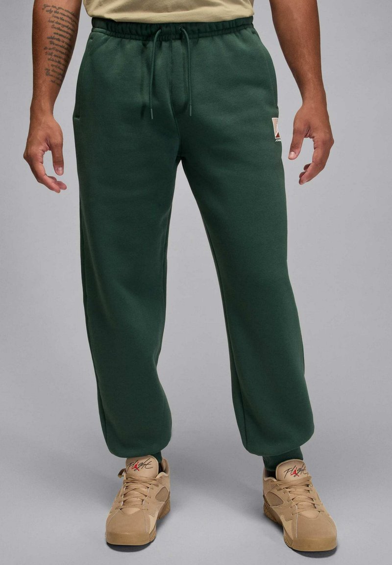 Grüne Jogginghose mit Tunnelzug in der Taille, zwei Seitentaschen und gerippten Bündchen. Kombiniert mit beigen Sneakers mit roten Akzenten.