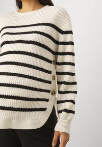 Knitted sweater with horizontal black and white stripes, round neckline, and side buttons. Textured fabric with a loose fit.