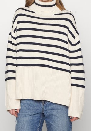 Personne portant un pull à col roulé crème avec des rayures noires horizontales associé à un jean bleu, debout devant un fond uni.