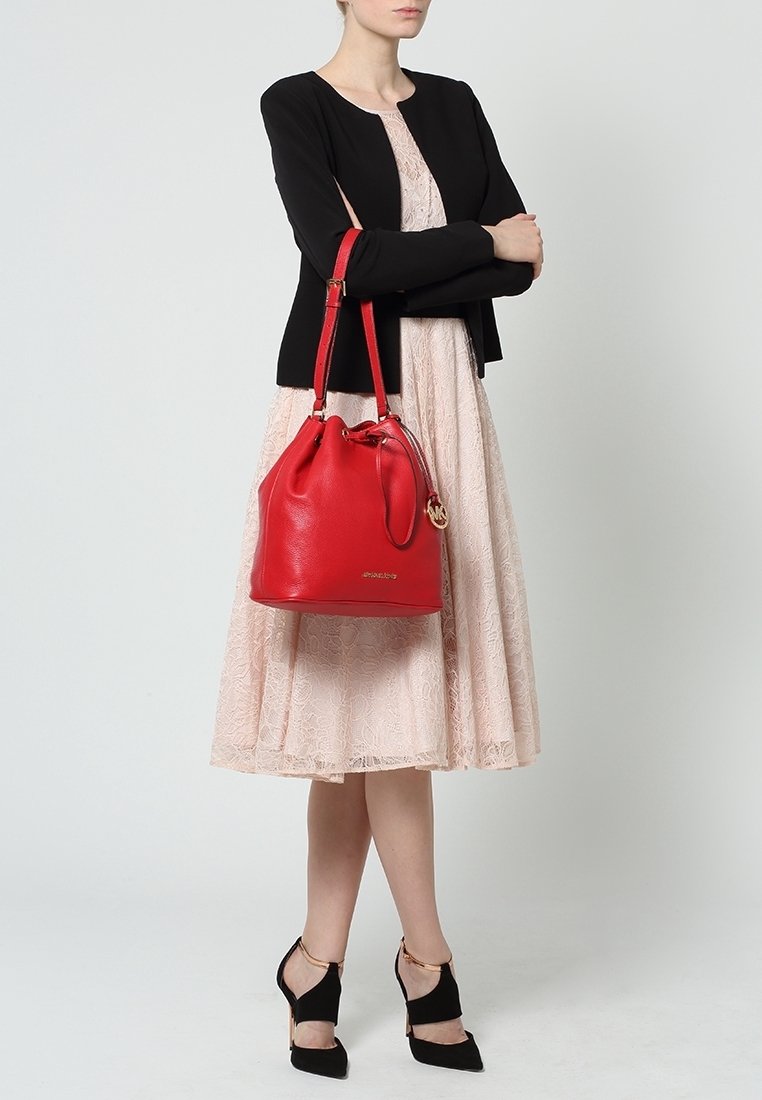 Femme en robe en dentelle rose pâle, veste courte noire, talons hauts noirs, tenant un grand sac à main en cuir rouge avec des bretelles d'épaule.