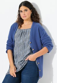 Cardigan blu lavorato a maglia con chiusura a bottone e polsini arrotolati, indossato sopra un top grigio a motivo con pois blu chiaro, abbinato a jeans scuri.