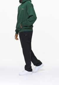 Vihreä huppari kengurun taskulla ja kirjailulla logolla, yhdistetty mustiin rennon istuvuuden housuihin ja valkoisiin tennareihin.