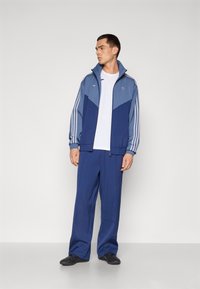 Training complet cu un design bleu deschis și bleu închis, având trei dungi albe pe mâneci, combinat cu pantaloni ușori și pantofi negri.