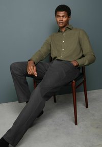Cămașă button-up de un verde măsliniu cu țesătură texturată, asortată cu pantaloni de un gri închis. Modelul este așezat pe un scaun din lemn, pe un fundal discret.