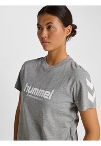 Graues T-Shirt mit kurzen Ärmeln und weißem "hummel"-Logo sowie Text. Verfügt über weiße Chevron-Muster an den Ärmeln. Hergestellt aus weichem, strukturiertem Stoff.