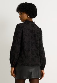 Blusa negra bordada con patrones florales, cuello alto y mangas largas con puños abotonados, complementada con una falda de cuero.