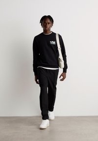 Zwart sweatshirt met een klein patroon op de zak, gecombineerd met zwarte broek en witte sneakers, en een lichtgekleurde schoudertas.