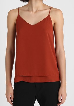 Femme portant un caraco sans manches couleur rouille à encolure en V et un pantalon noir, sur un fond clair uni.