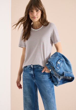 Jeune femme portant un t-shirt à rayures blanches et marron et un jean bleu, tenant une veste en denim bleue pliée dans sa main gauche.