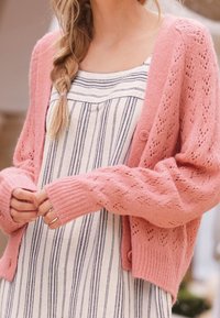 Žena s pletenými blond vlasy, která nosí růžový pletený cardigan přes bílou šaty s vertikálními černými pruhy, upravuje rukávy cardigan.