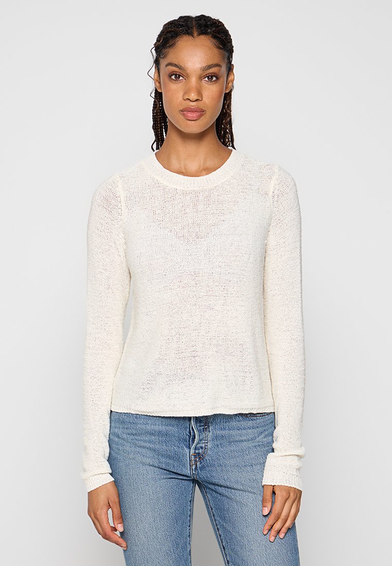 Kremowy sweter z długim rękawem, wykonany z teksturowanej dzianiny, z okrągłym dekoltem. Noszony z niebieskimi dżinsami, z dopasowanymi rękawami.